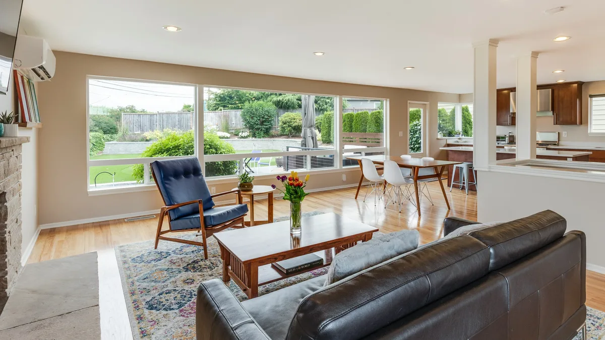 A living room with a brown leather couch, a blue chair, a coffee table, and a rug on a hardwood floor, with a kitchen visible in the background and a large window offering a view of the outdoors.